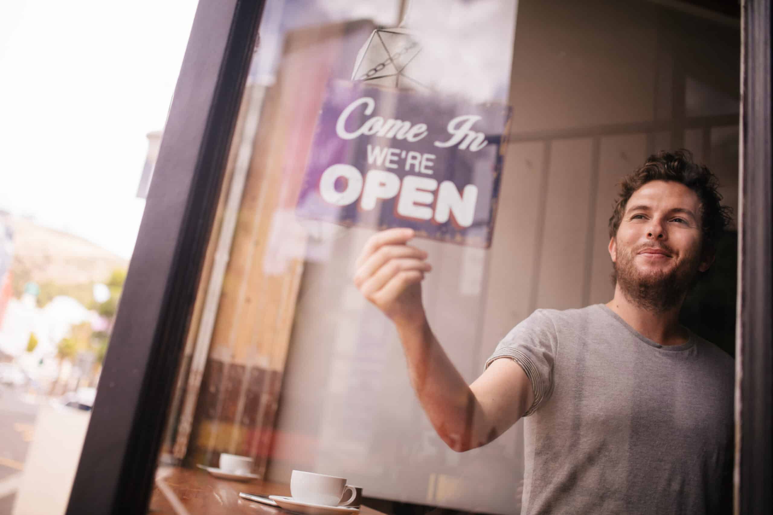 Man hanging an 'We're Open' sign in a cafe window.