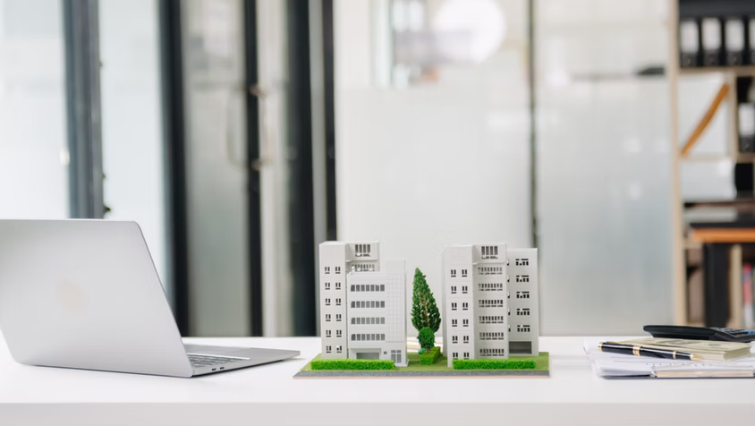 Architectural model of buildings and trees on a white desk with a laptop, calculator, pen, and cash.