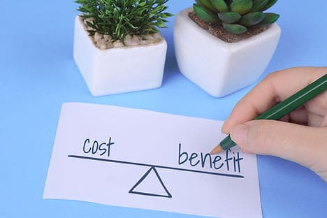 Hand writing 'cost benefit' on paper with plants in background.