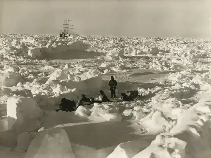 Researchers navigating icy Antarctic landscape with ship in background.