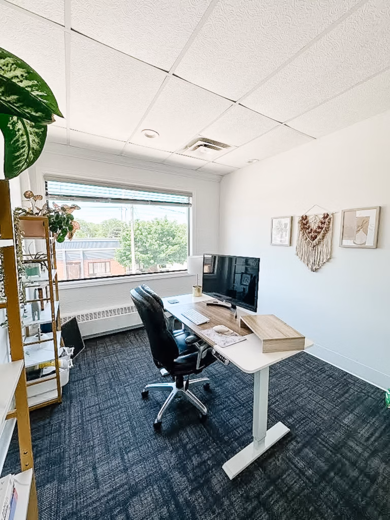 Modern office with standing desk, ergonomic chair, plants, window view, and patterned ceiling.