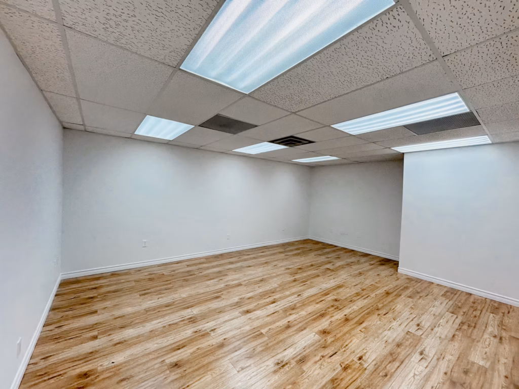 Empty commercial office space with white walls, wood-look flooring, and fluorescent lighting.