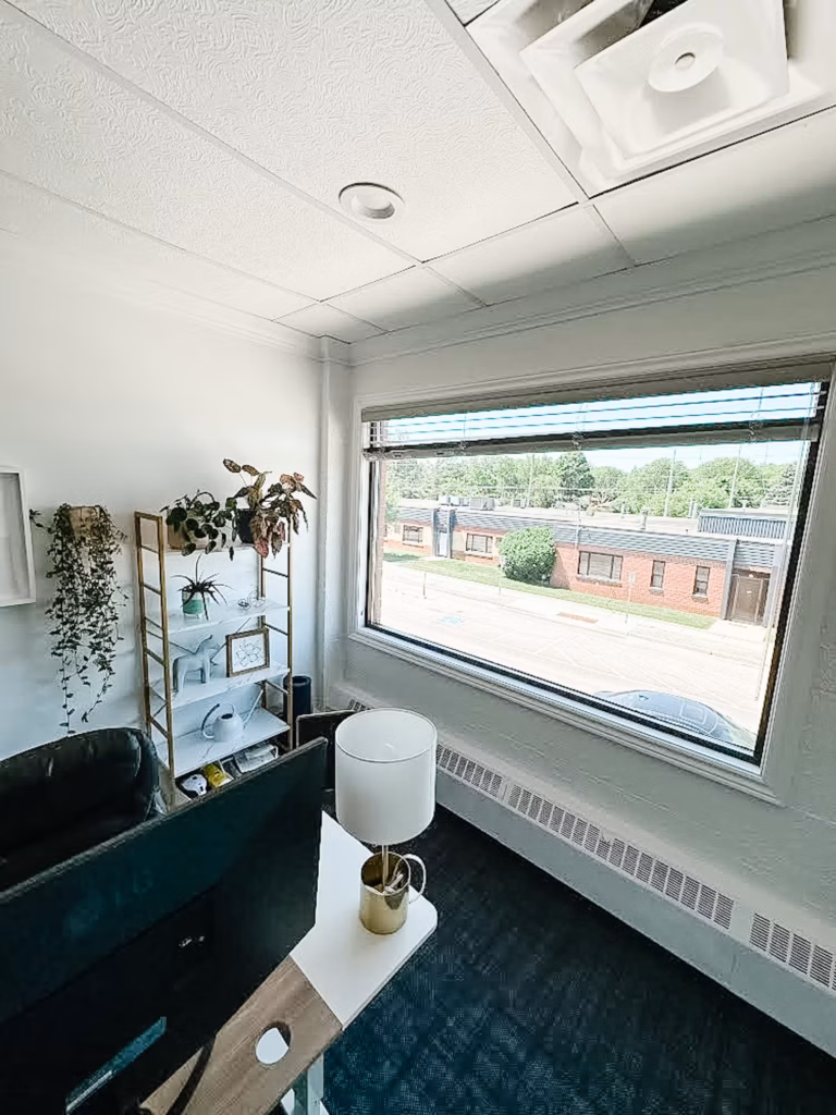 Modern office with plants, white desk, monitor, lamp, and window view of a brick building.