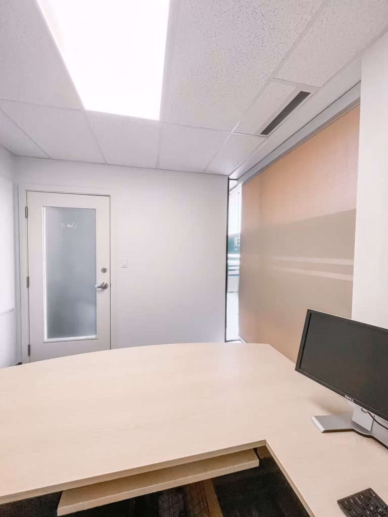 Bright office with light wood desk, monitor, frosted glass door "Suite 12", and window with blinds.