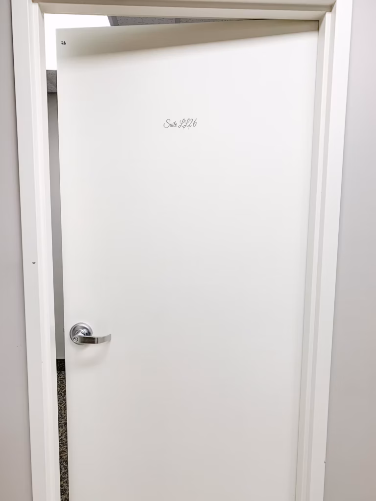 White door slightly ajar, revealing "26" and "Suite LL26" in black text. Silver doorknob.
