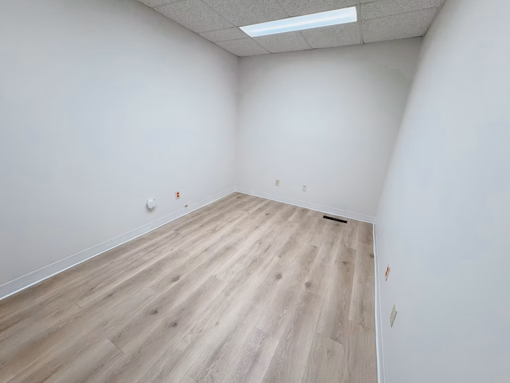 Empty office room with light wood flooring, white walls, and drop ceiling.