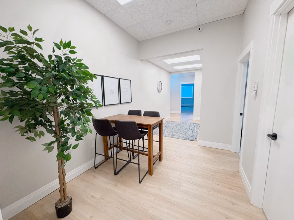 Modern office with a tall faux plant, high-top table, chairs, framed art, and light wood flooring.