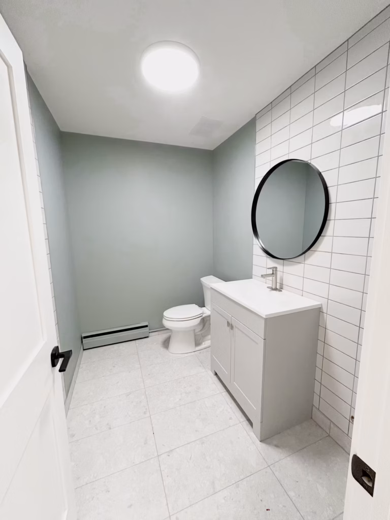 Modern bathroom with sage green walls, white subway tile, light gray vanity, and round mirror.