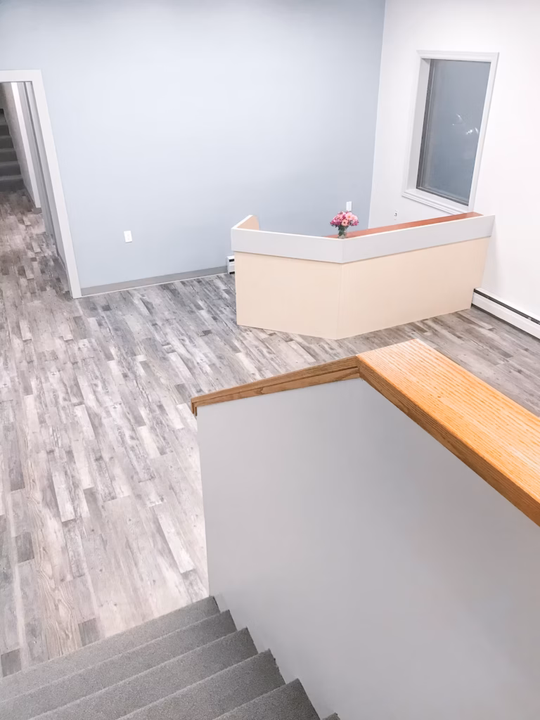 Modern reception area with light blue walls, wood-look floor, beige desk, and gray carpeted stairs.