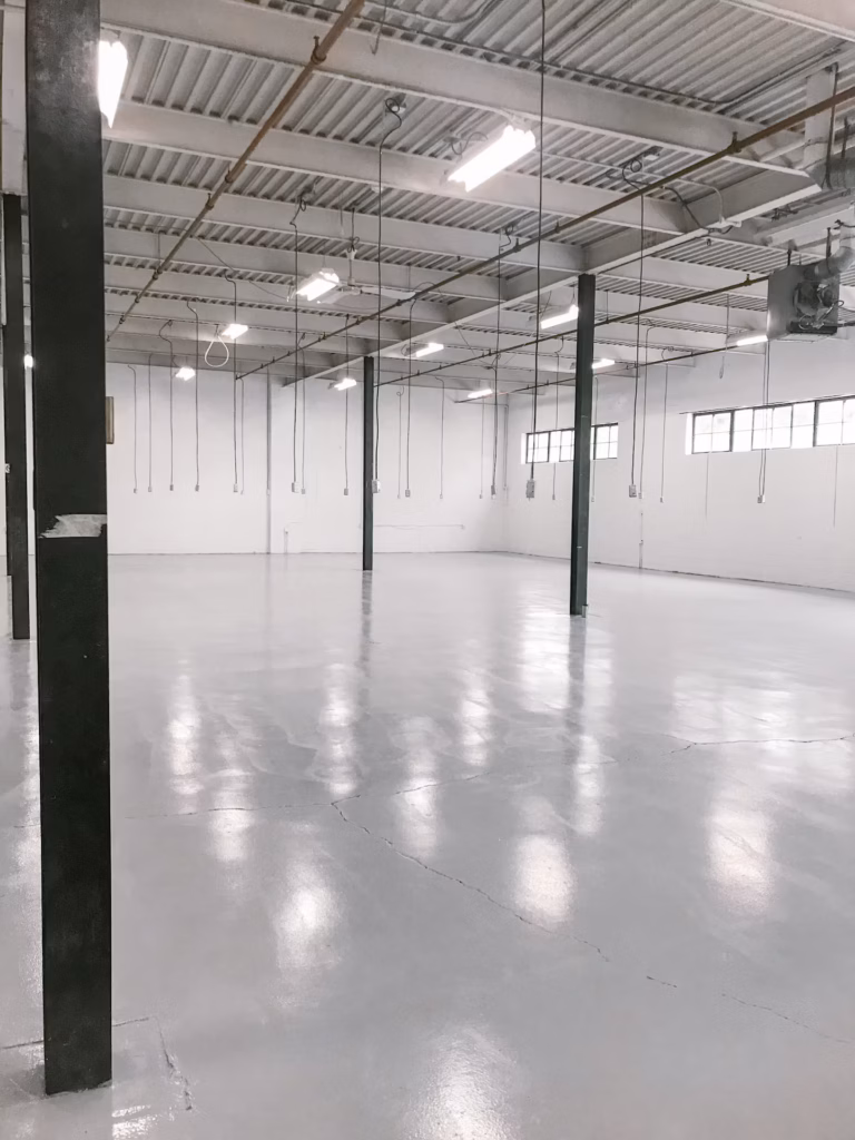 Empty, bright industrial space with white walls, grey floor, exposed ceiling, and windows.