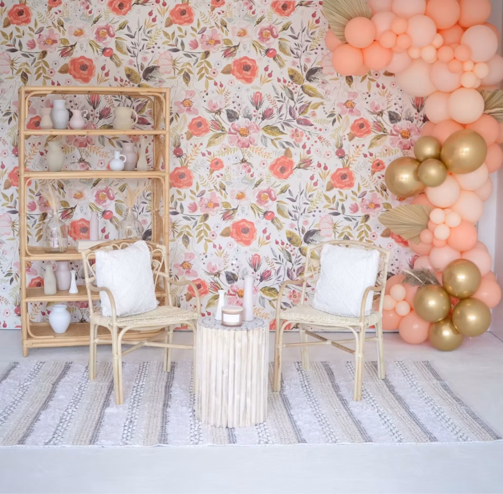 Boho nursery with floral wallpaper, rattan shelf, chair, table, rug, and peach & gold balloon arch.