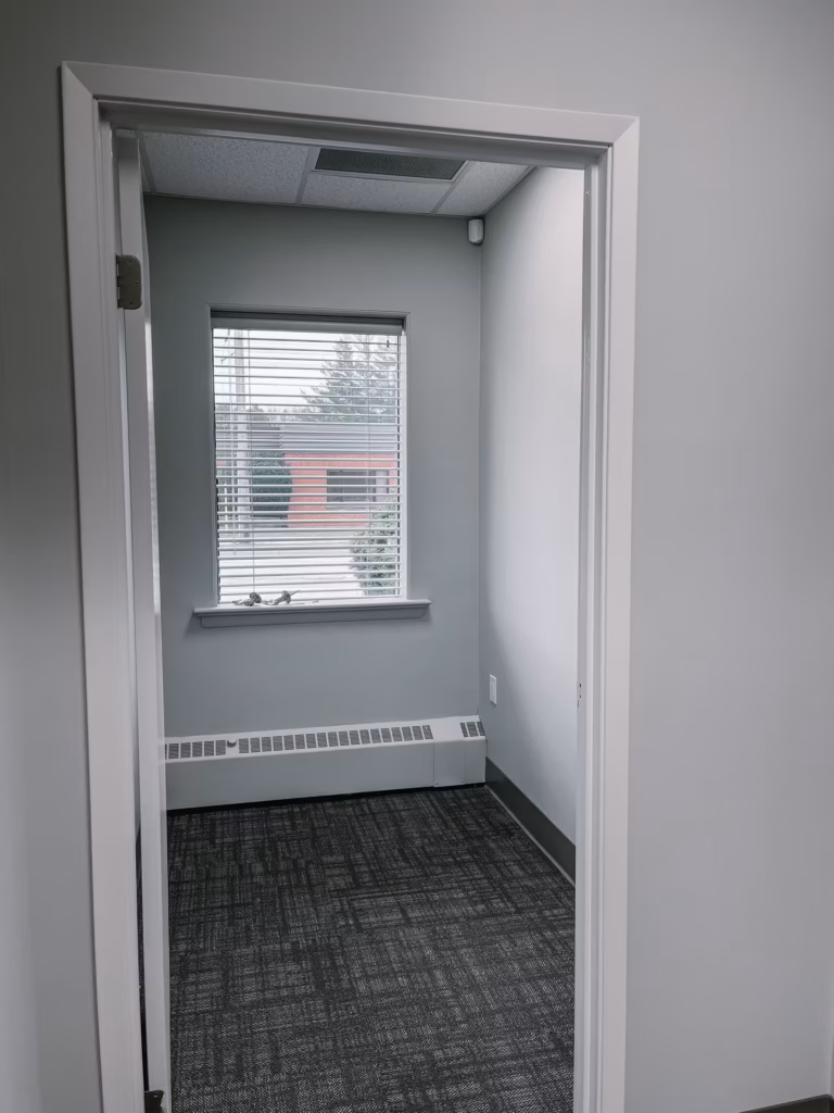 Empty office room with gray walls, dark carpet, window with blinds, and baseboard heater.