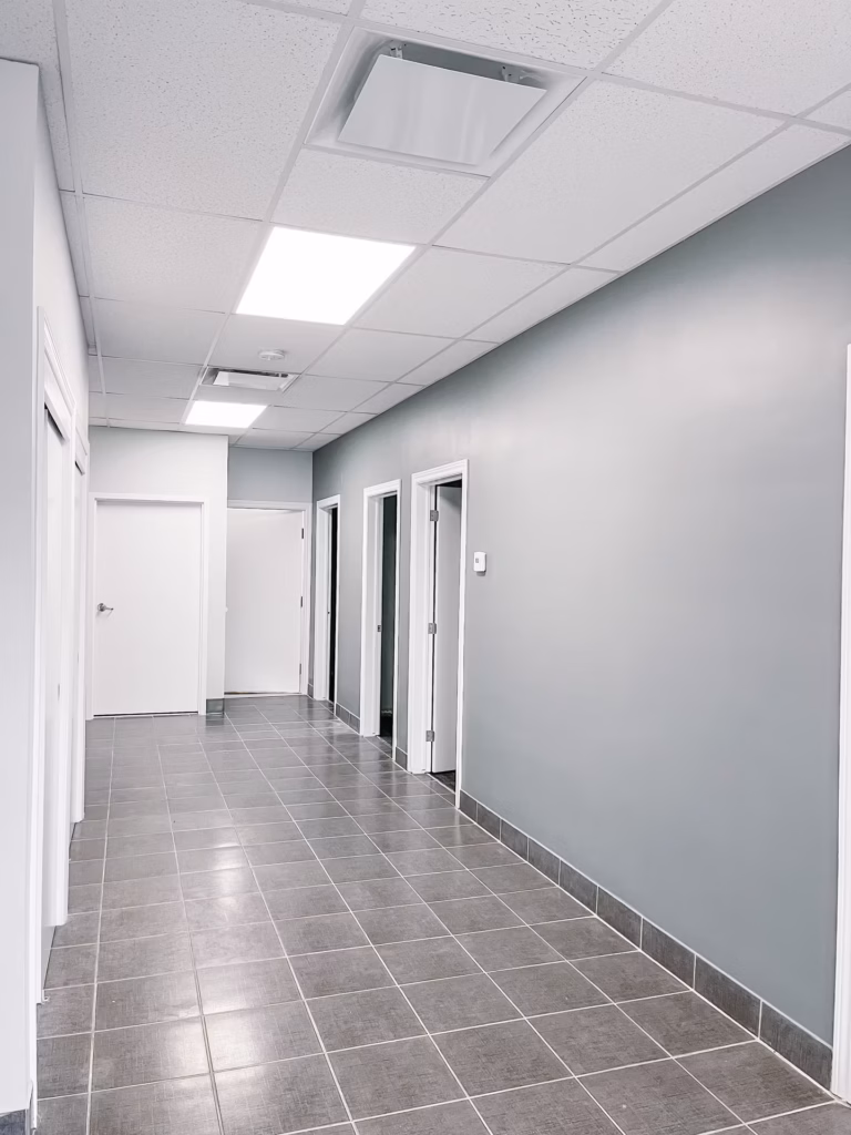 Modern office hallway with light gray walls, white doors, gray tiled floor, and bright ceiling lights.