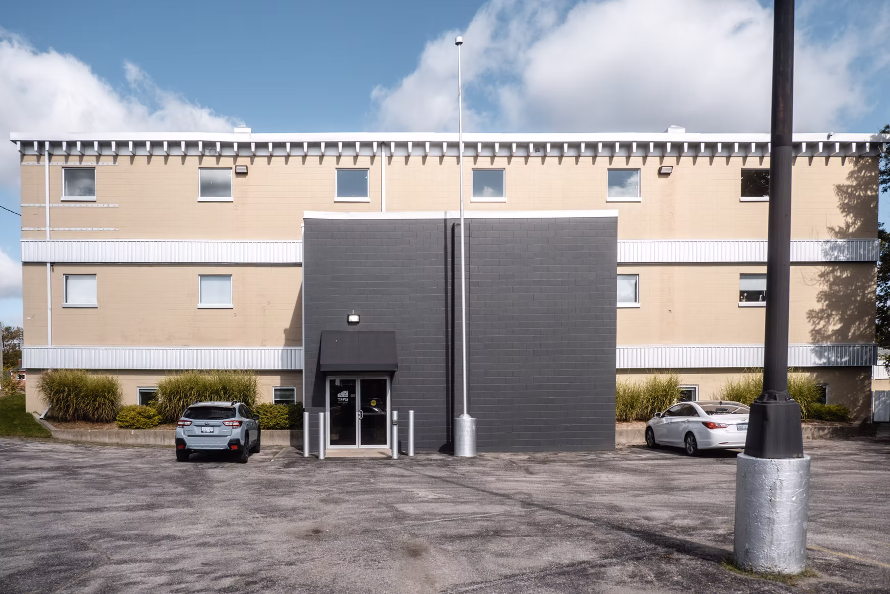 Building with tan walls, gray entrance, and parking lot. Cars parked. "TFPQ" on door.