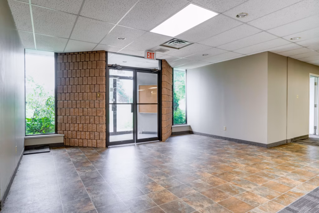 Empty office space with tile floor, brick wall, EXIT sign, and large windows looking outside.
