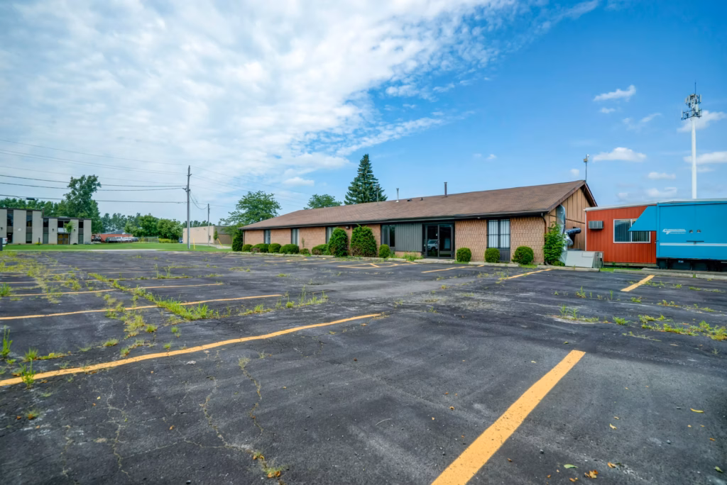 Dilapidated parking lot with weeds, office building, cell tower, and storage containers under blue sky.