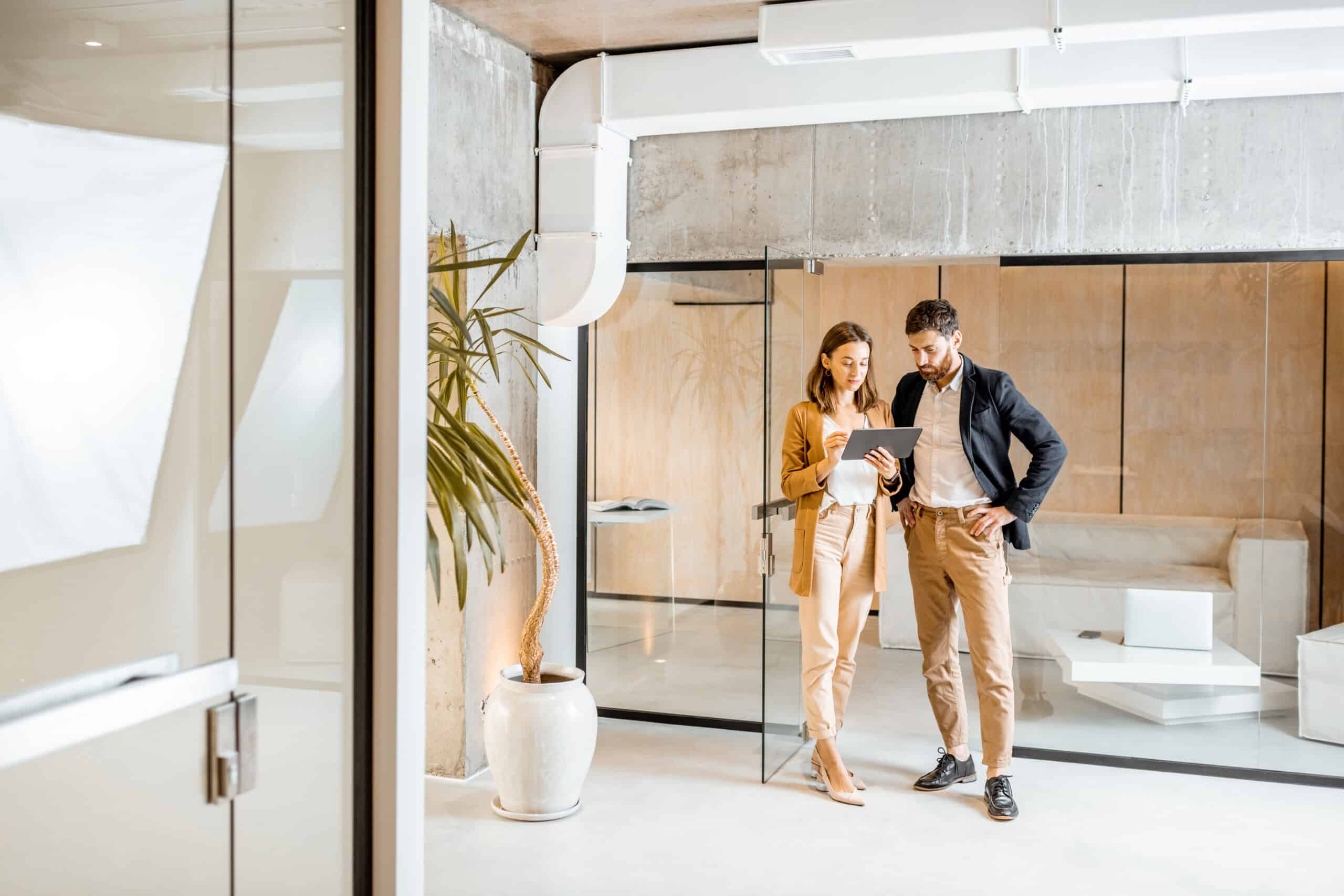 Two professionals discuss on a tablet in a modern office with glass walls and concrete ceiling.