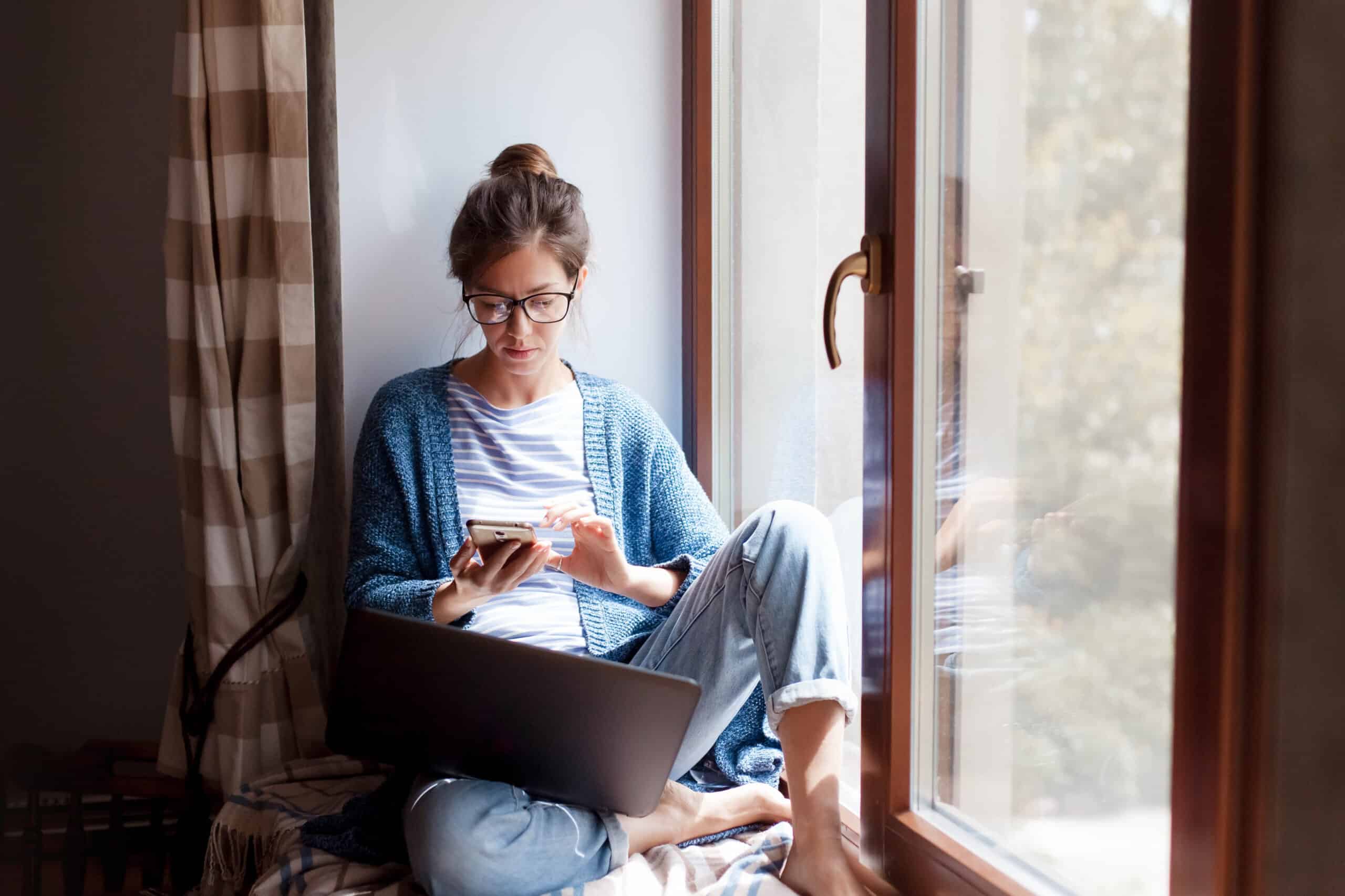 Woman working on laptop by window in home office setting.